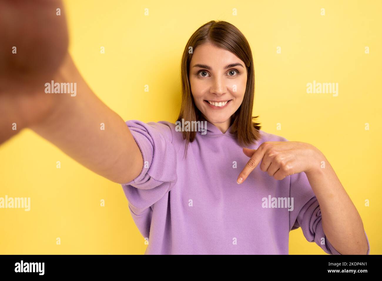 Souriante femme attrayante prenant selfie, point de vue de la photo, pointant le doigt vers le bas, recommander de souscrire son vlog, portant le pull à capuche violet. Studio d'intérieur isolé sur fond jaune. Banque D'Images