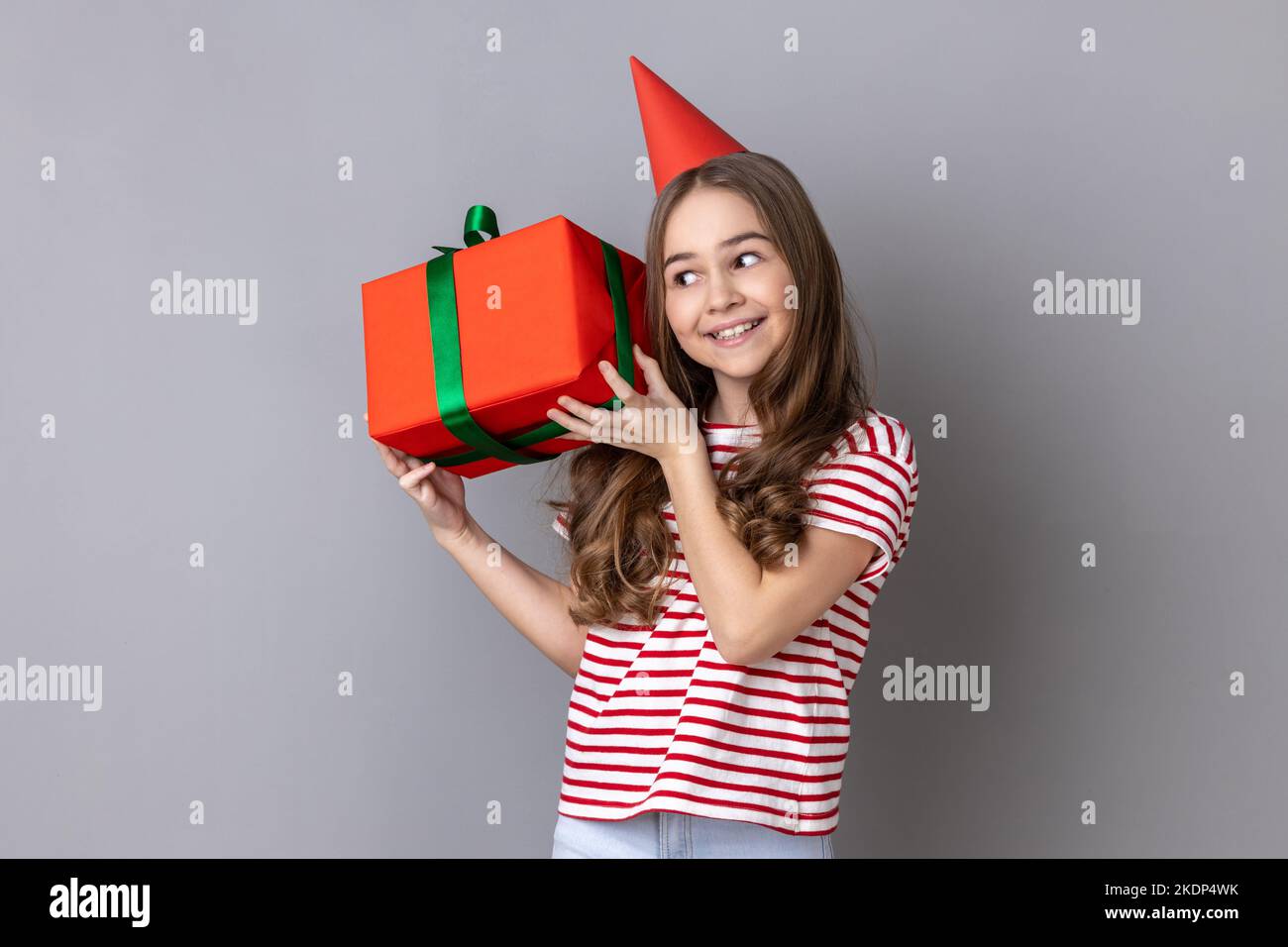 Portrait d'une petite fille portant un T-shirt rayé et un cône de fête tenant une boîte cadeau enveloppée de bleu, étant intéressé blanc à l'intérieur du cadeau. Prise de vue en studio isolée sur fond gris. Banque D'Images