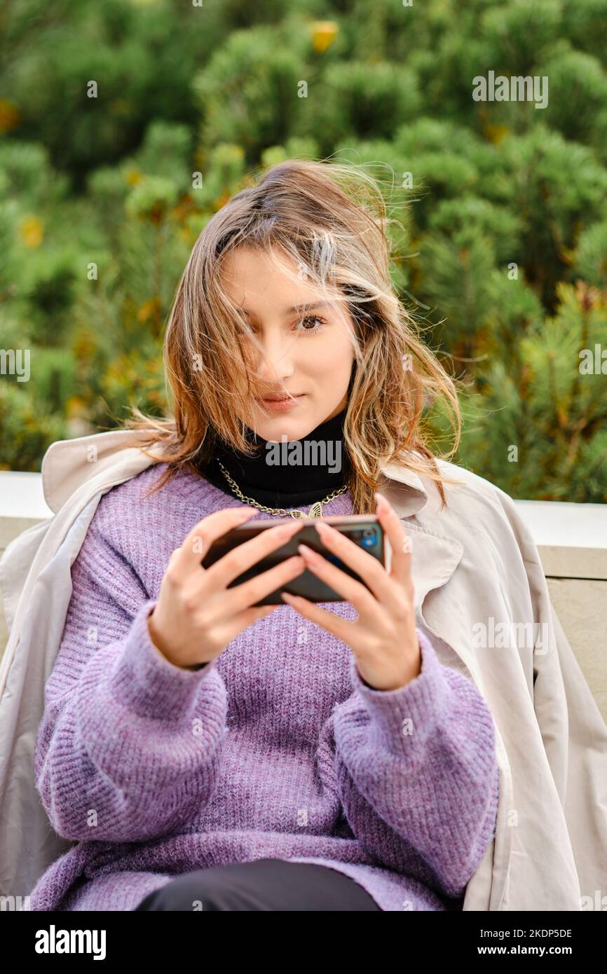 Une jeune femme est assise sur un banc et joue à un jeu ou regarde une vidéo sur un smartphone Banque D'Images