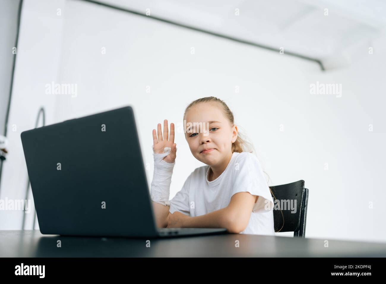 Positive petite fille avec cassé main enveloppée dans le bandage en plâtre blanc montrant bonjour geste regardant la caméra assis au bureau avec ordinateur portable dans la lumière Banque D'Images