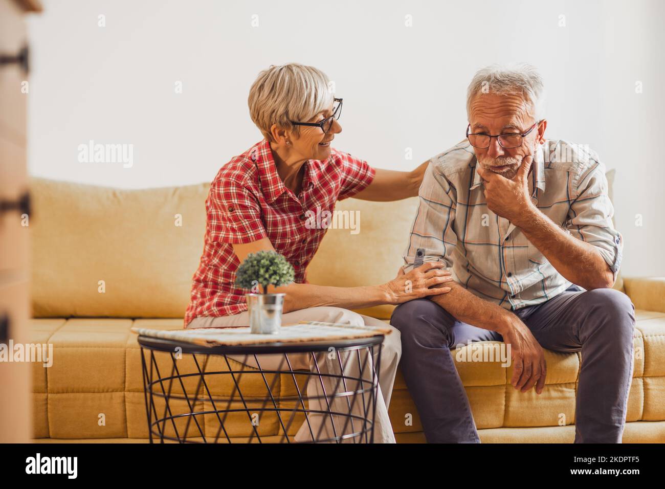 Couple senior assis dans la salle de séjour. L'homme est triste et inquiet et la femme lui console. Banque D'Images