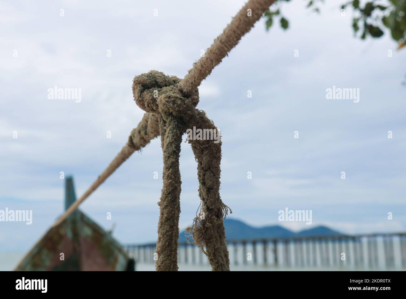 corde brune ayant un nœud sur une corde reliée à un bateau en bois par la plage avec vue sur la plage, jetée en bois avec île et ciel nuageux Banque D'Images