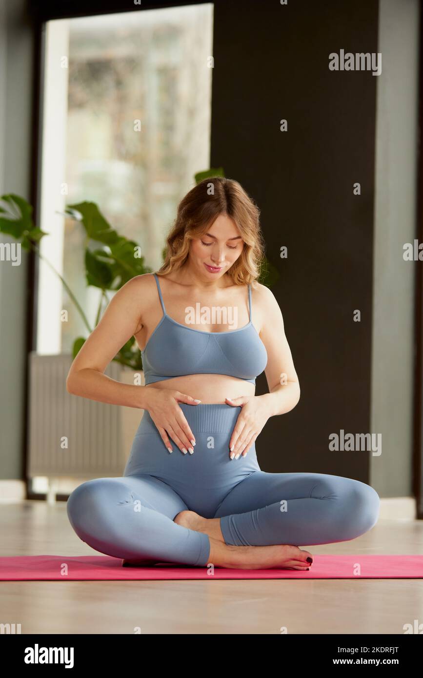 Portrait de la jeune femme enceinte dans des vêtements de sport assis sur un tapis de yoga et tenant le ventre avec la forme de coeur. Banque D'Images