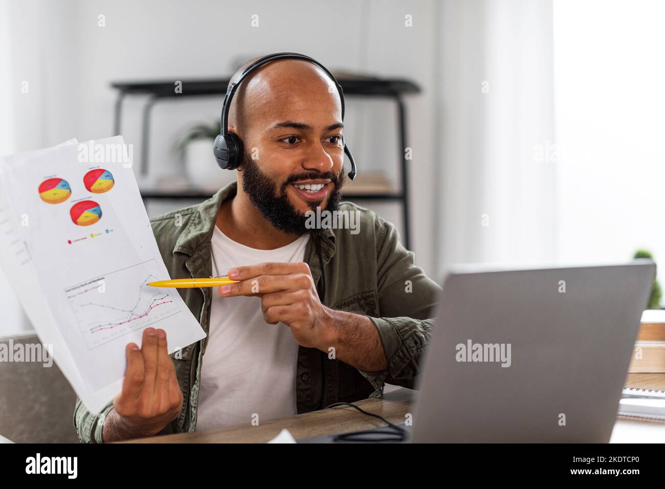 Homme latin mûr souriant dans un casque montrant des graphiques à la webcam d'ordinateur portable, assis au bureau à la maison, espace libre Banque D'Images
