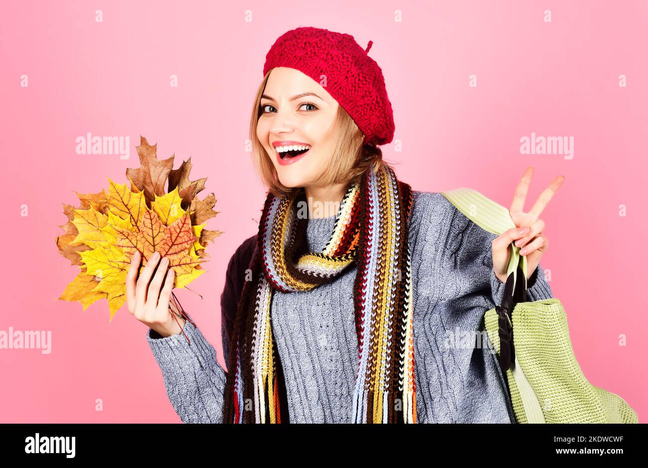 Femme souriante avec sac à provisions et feuilles jaunes dans le chandail, chapeau et foulard montre la paix signe. Banque D'Images