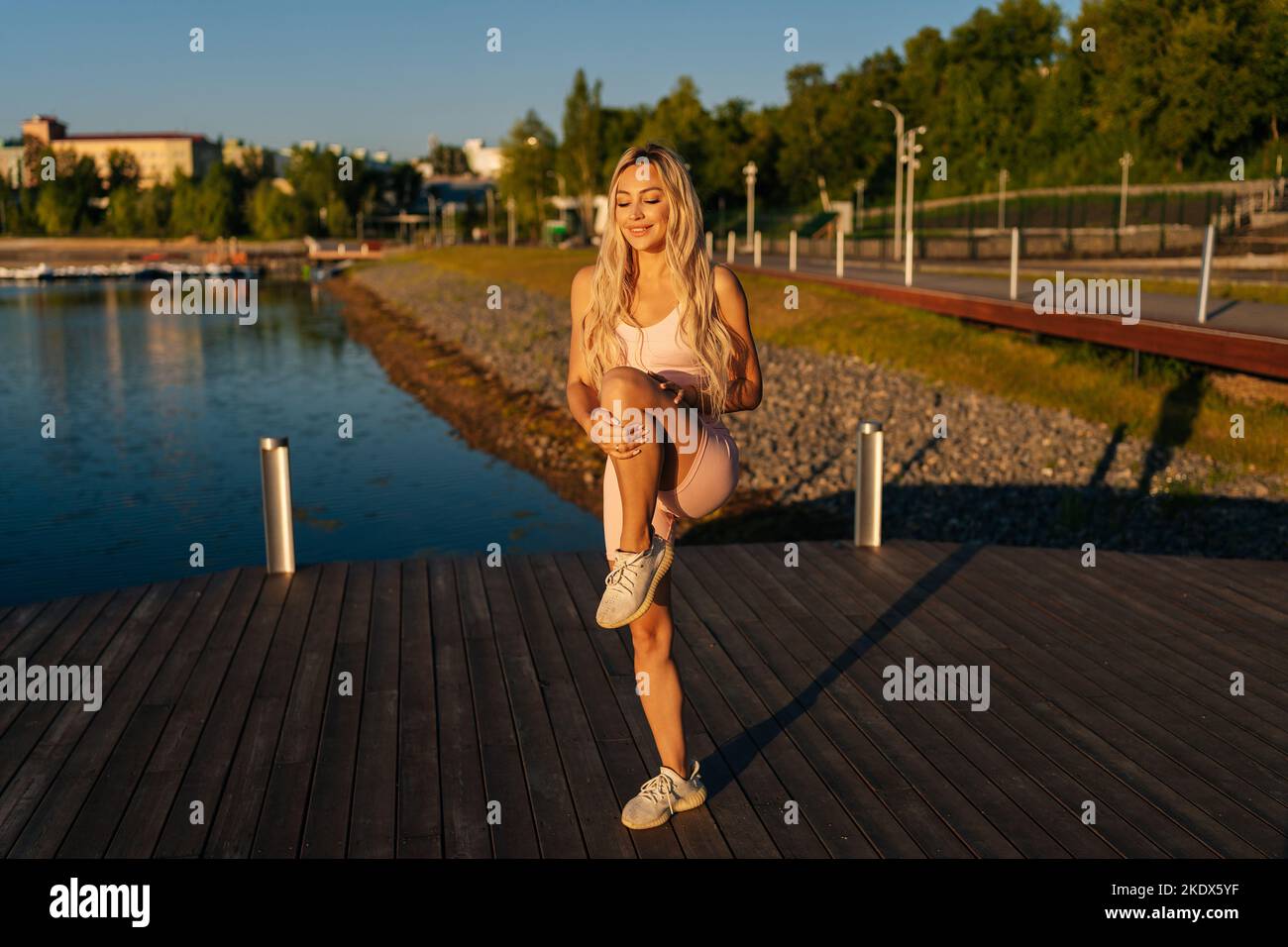 Jeune femme blonde souriante se préparant à l'entraînement cardio, étirant les muscles des jambes, tenant le genou à la poitrine debout près de l'eau dans le parc de la ville. Banque D'Images