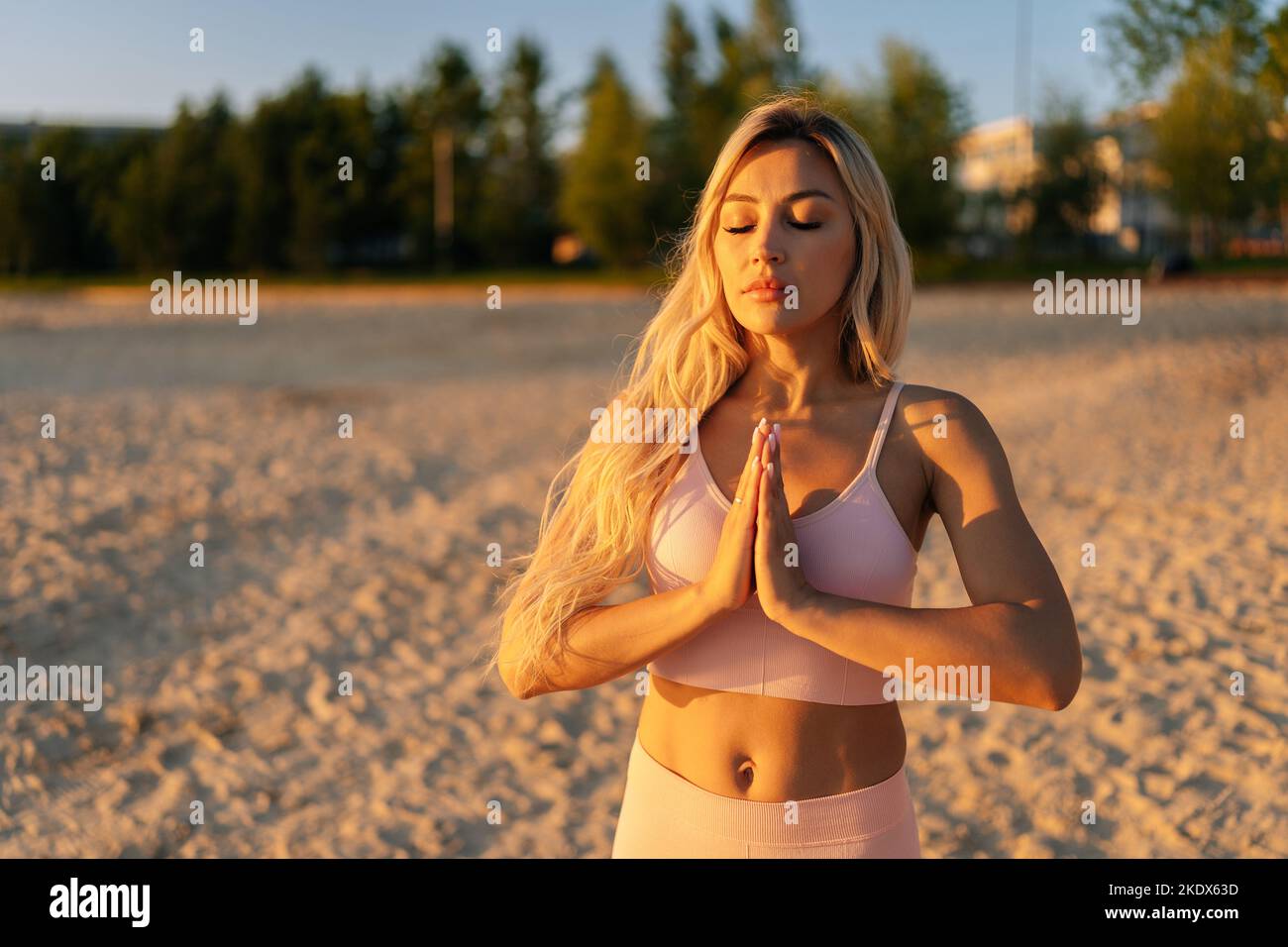 Portrait de jeune femme calme avec les yeux fermés tenant les mains dans un geste namaste debout sur la plage de sable à l'été matin ensoleillé. Banque D'Images