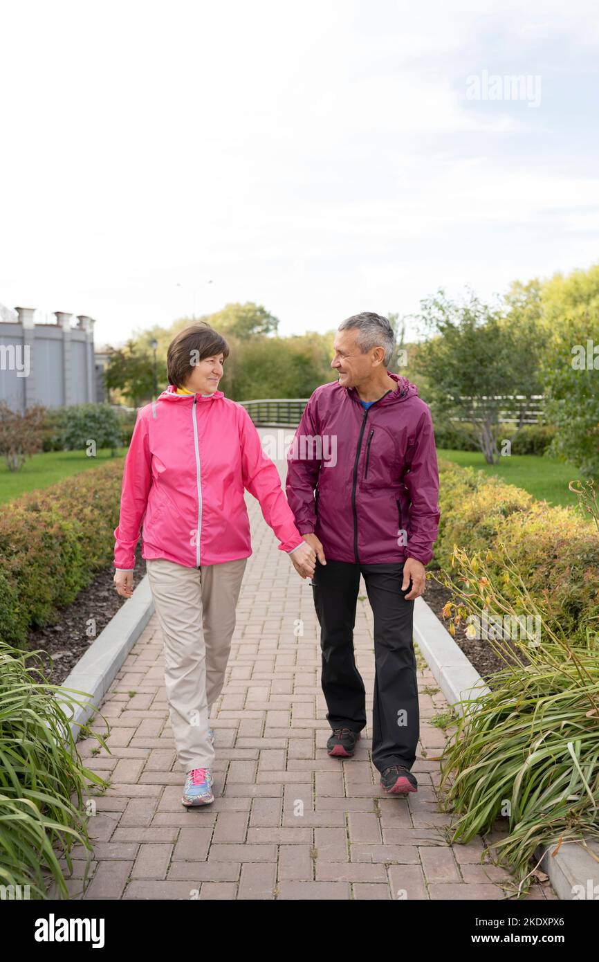 Tout le corps heureux homme et femme d'âge moyen tenant les mains et regardant l'un l'autre tout en se promenant sur le chemin pavé dans le parc Banque D'Images