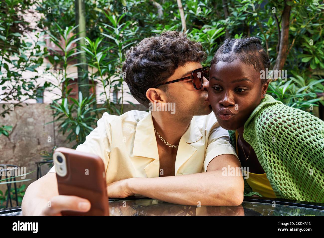 Un jeune homme élégant embrasse une petite amie noire qui se repose sur une table et prend son selfie via un smartphone tout en étant assise à table pendant un séjour romantique Banque D'Images