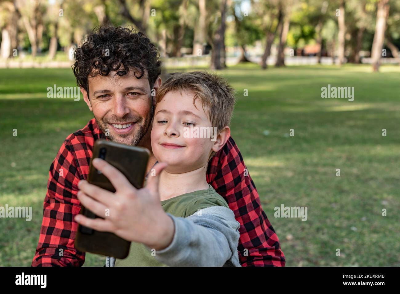 Un fils positif qui se fait autoportrait sur un smartphone avec un papa heureux tout en étant assis sur l'herbe en forêt en été Banque D'Images