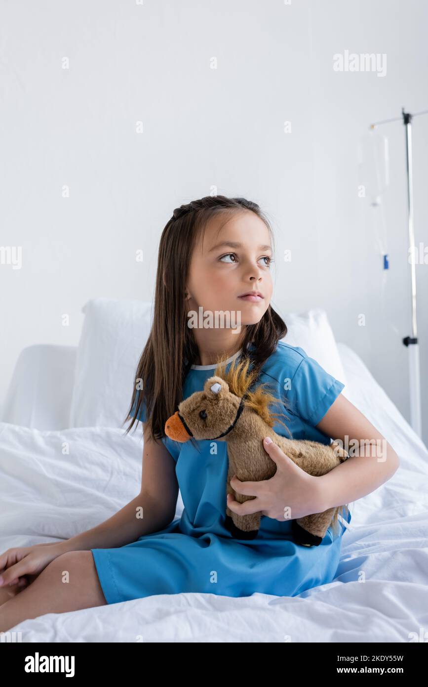 Enfant dans la robe de patient tenant le jouet tout en étant assis sur le lit dans la clinique, image de stock Banque D'Images