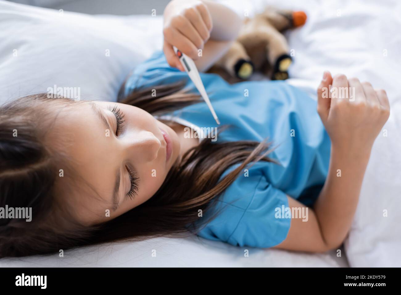 Fille dans la robe de patient tenant thermomètre électronique flou sur le lit dans la clinique, image de stock Banque D'Images