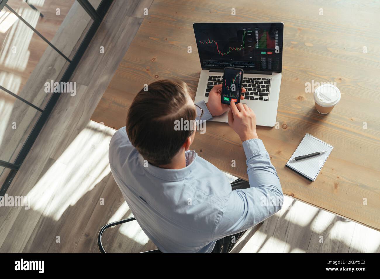 Vue de dessus d'un homme assis à une table de bureau devant un ordinateur portable avec des cartes du marché de la crypto-monnaie, tenant un téléphone avec une application d'investissement ouverte avec un diagramme de prix allant appuyer sur le bouton de vente. Copier l'espace Banque D'Images