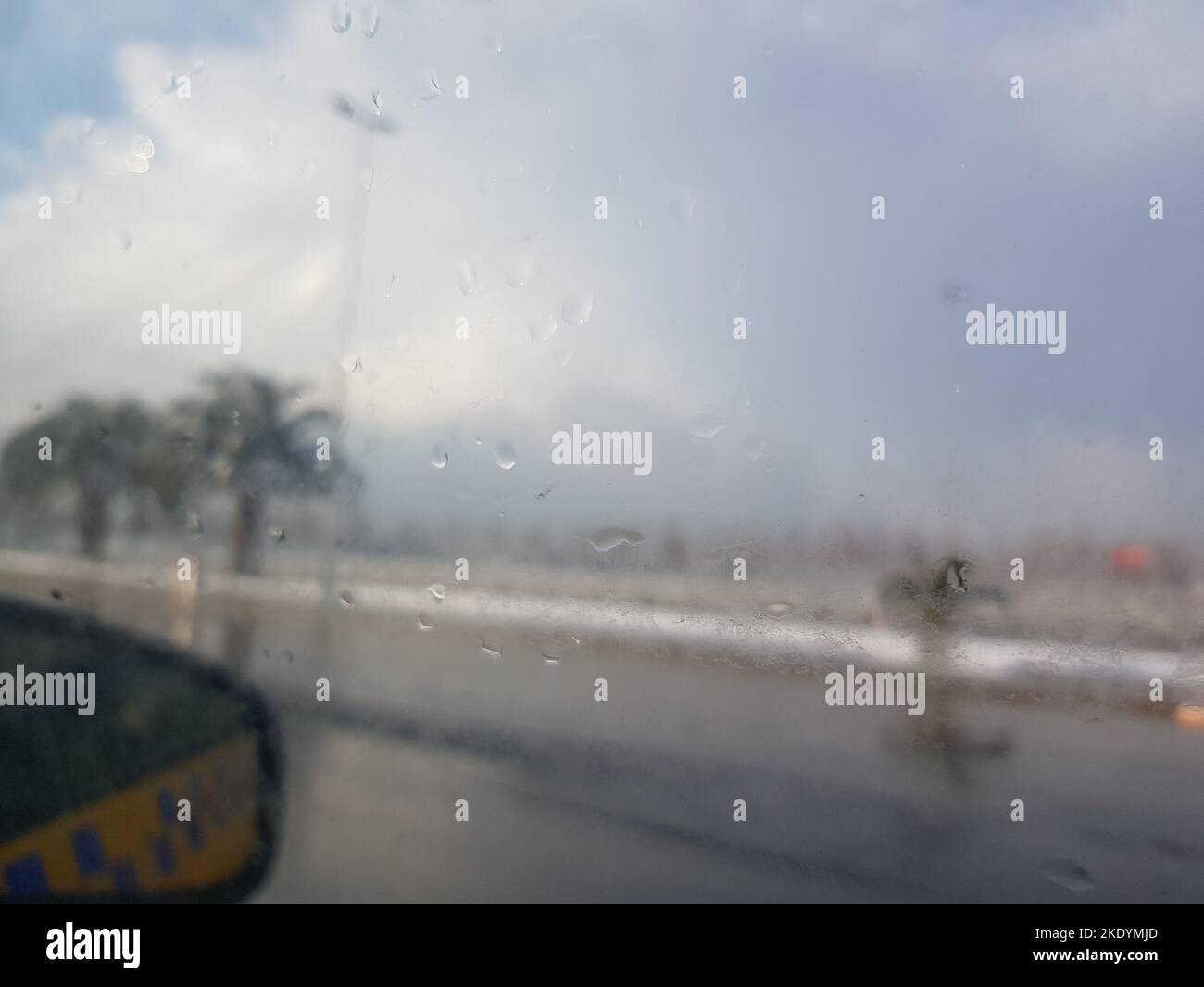 La pluie tombe sur la fenêtre de la voiture avec une vue du miroir extérieur et un arrière-plan flou avec des palmiers Banque D'Images