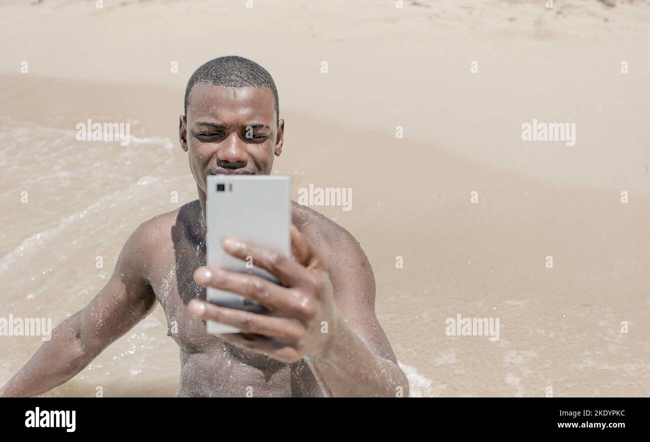 Vue de face Noir homme voyageur assis sur le sable près de la mer et prenant un selfie pendant les vacances d'été à la station. Banque D'Images