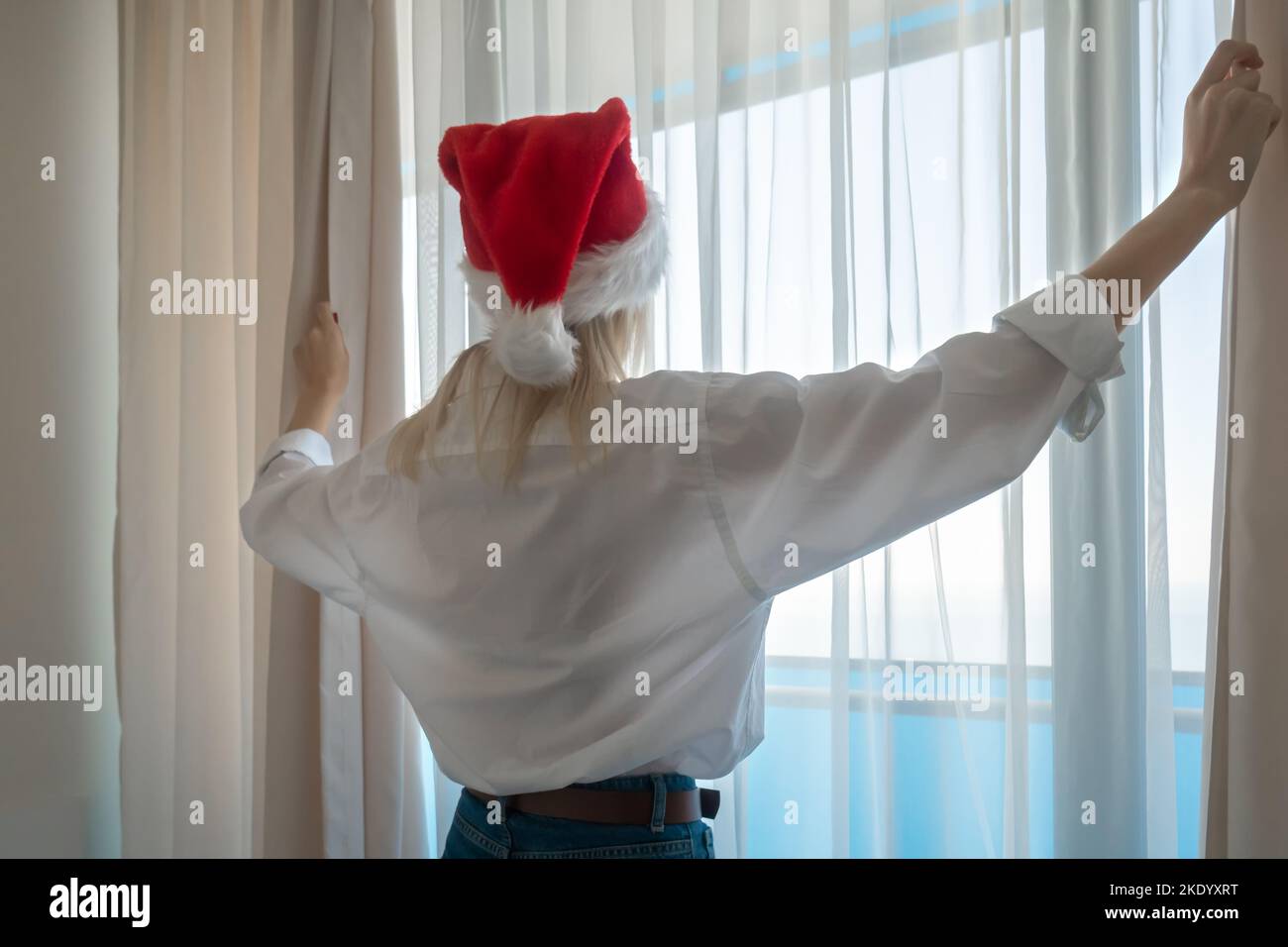 Gros plan d'une jeune femme dans un chapeau de Père Noël dans une chemise blanche et jeans bleus poussant les rideaux, photo de côté Banque D'Images