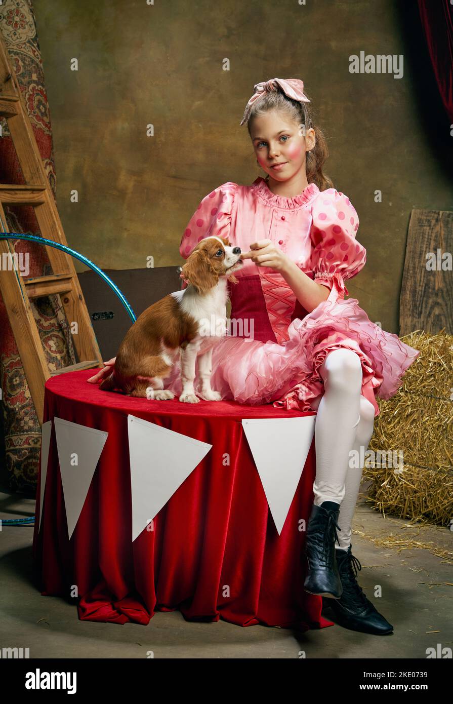 Entraîneur de cirque. Portrait de la petite jolie fille dans la tenue de fête formation drôle de chien au cirque vintage. Vacances, rêves, art, mode Banque D'Images