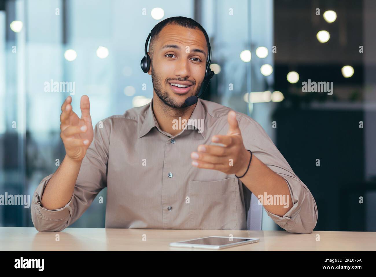 Vue webcam, heureux afro-américain homme parlant en ligne appel vidéo regardant la caméra à l'aide d'un casque, homme travaillant à l'intérieur de l'assistance technique moderne de bâtiment de bureau et centre d'appel d'assistance téléphonique. Banque D'Images