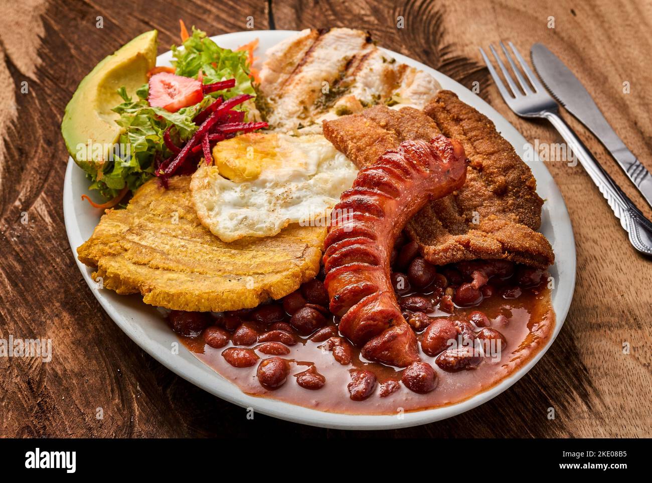 Vue de dessus d'une assiette blanche avec Bandeja paisa avec un œuf, des saucisses et une salade de légumes sur une table en bois Banque D'Images