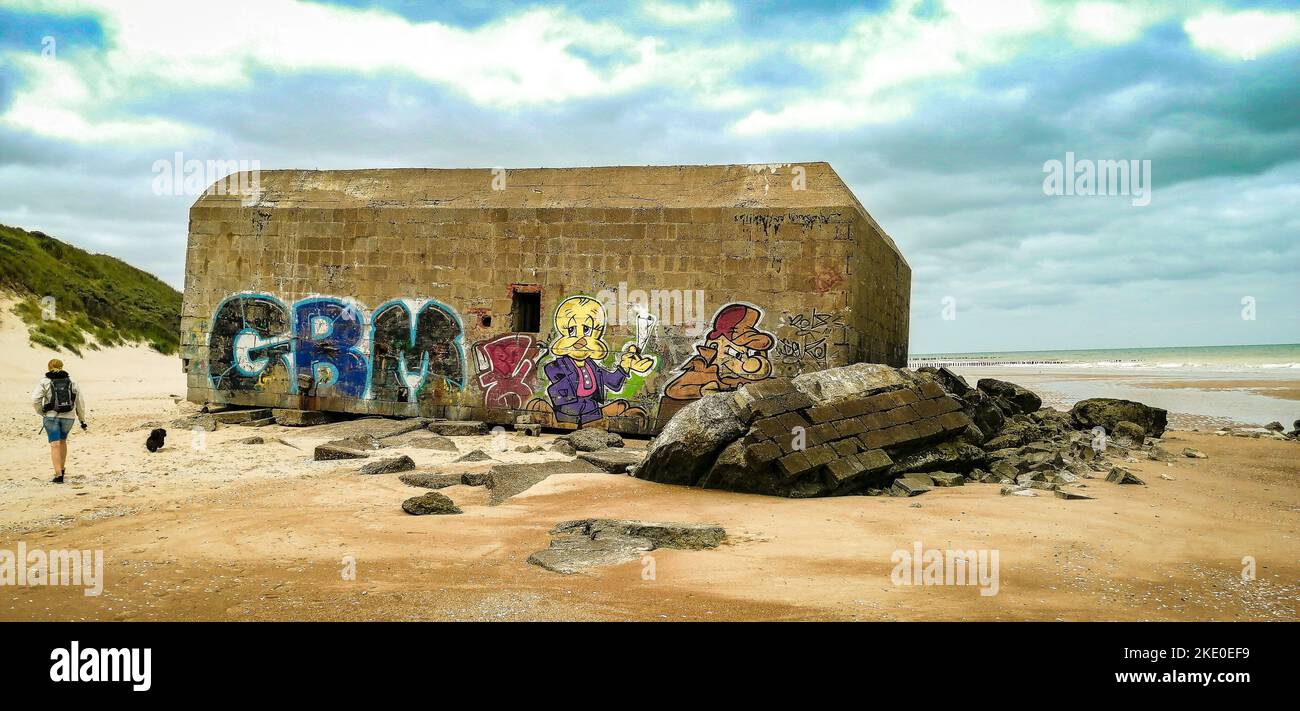 Un homme et un chien marchant sur une plage en France à côté des ruines de bunker avec des graffitis de dessins animés colorés Banque D'Images