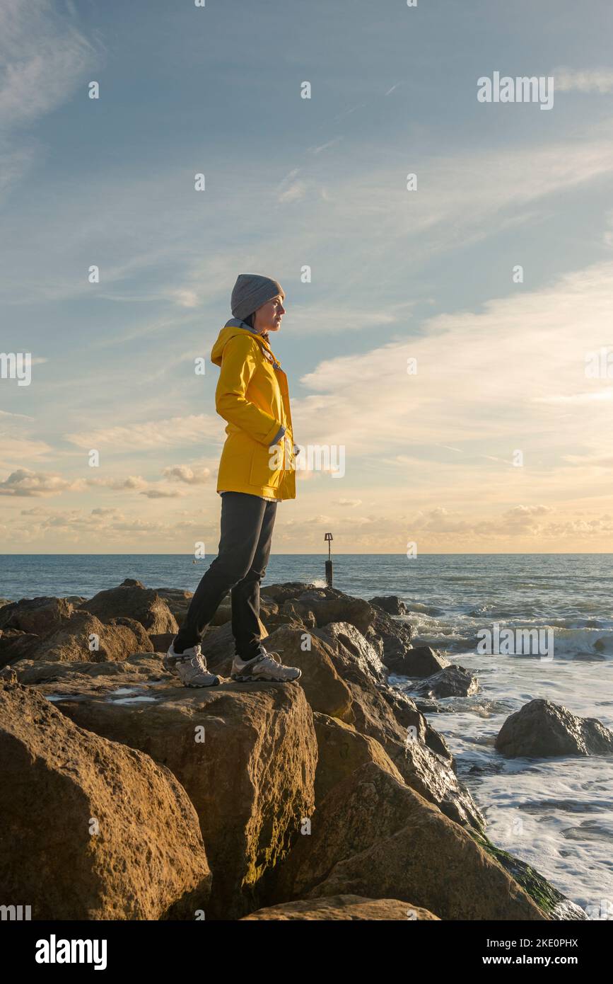 Femme debout sur des rochers regardant l'océan, manteau jaune. Banque D'Images