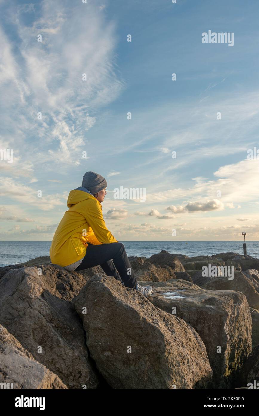 Femme assise sur des rochers au bord de l'océan, regardant les vagues. Banque D'Images