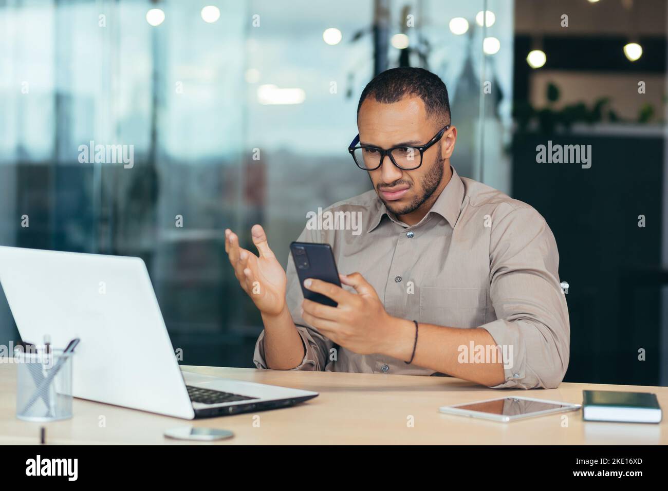 Un homme afro-américain a reçu de mauvaises nouvelles en ligne du téléphone, homme d'affaires en chemise travaillant avec un ordinateur portable au milieu d'un immeuble de bureau moderne, homme en lunettes lisant une lettre en ligne. Banque D'Images