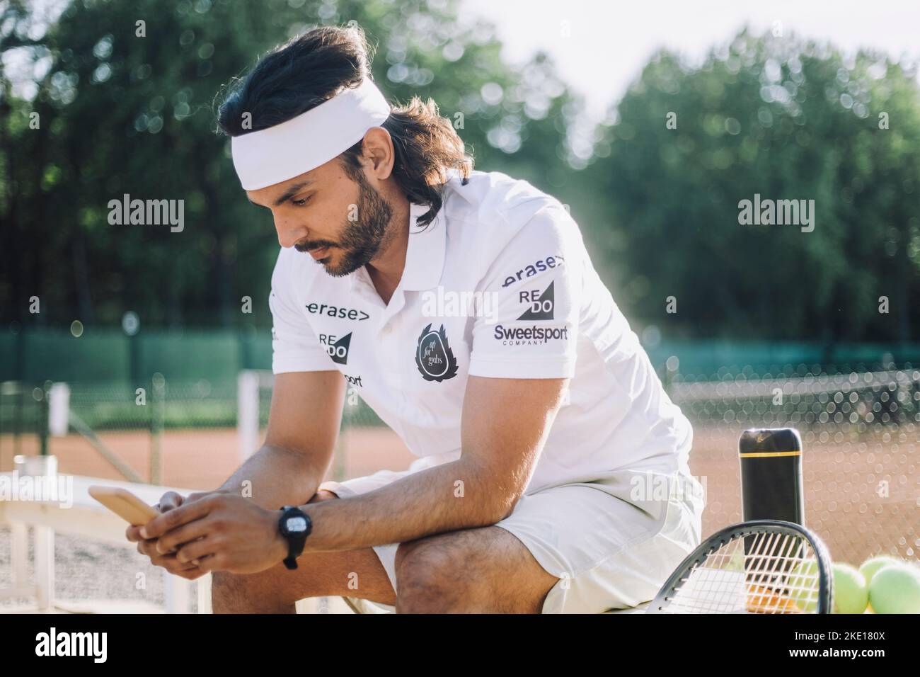 Homme portant un serre-tête à l'aide d'un smartphone assis sur un banc sur un court de tennis Banque D'Images