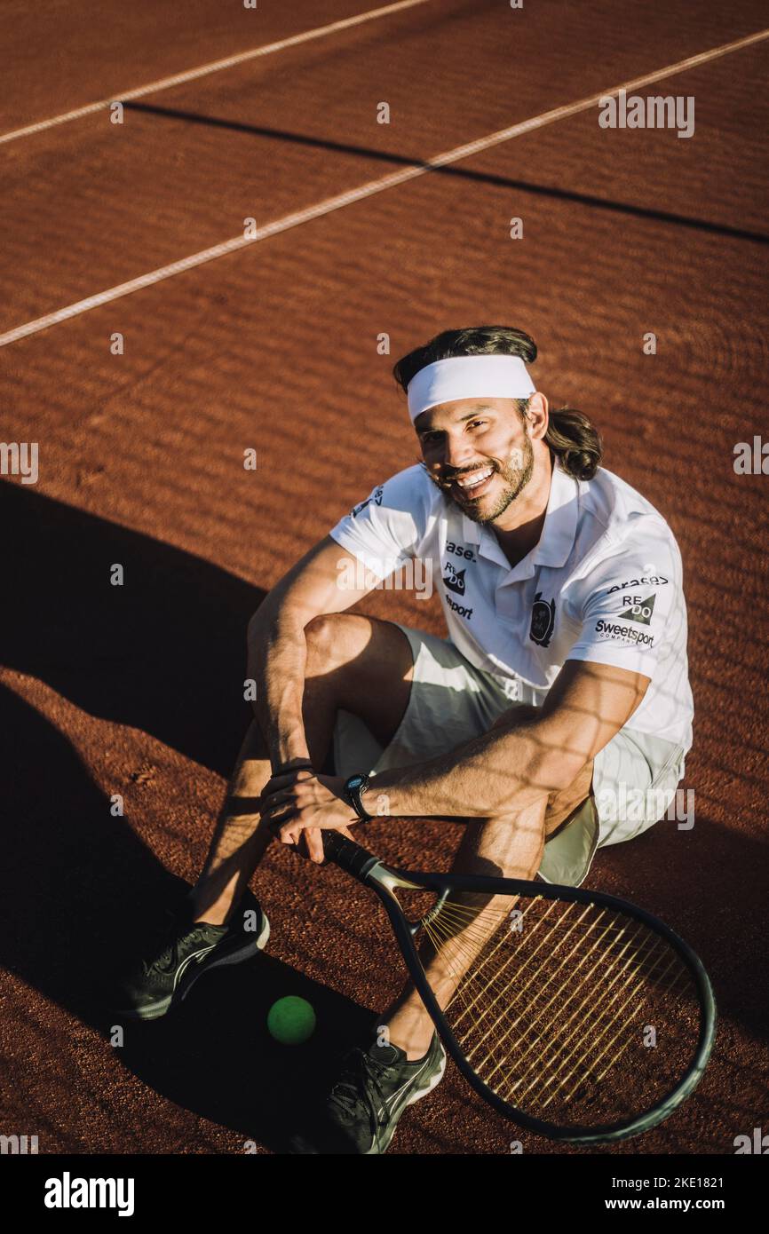 Vue en grand angle de l'homme souriant avec une raquette de tennis assise sur le sol pendant la pause Banque D'Images