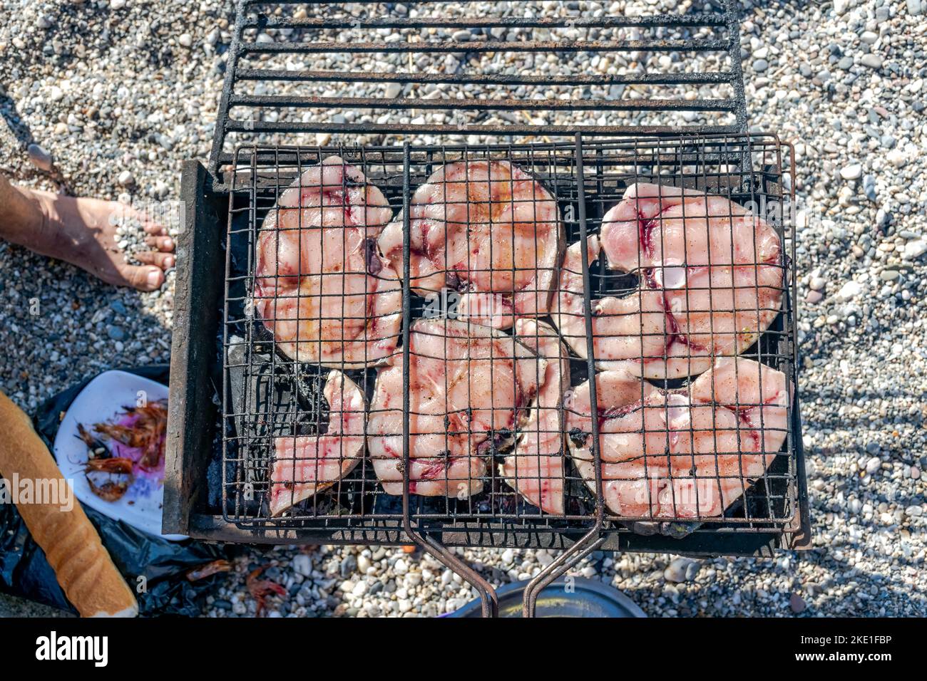 Vue aérienne de tranches d'espadon grillées sur un barbecue, un pain, une assiette de crevettes et un pied avec des galets de plage sur fond flou. Banque D'Images