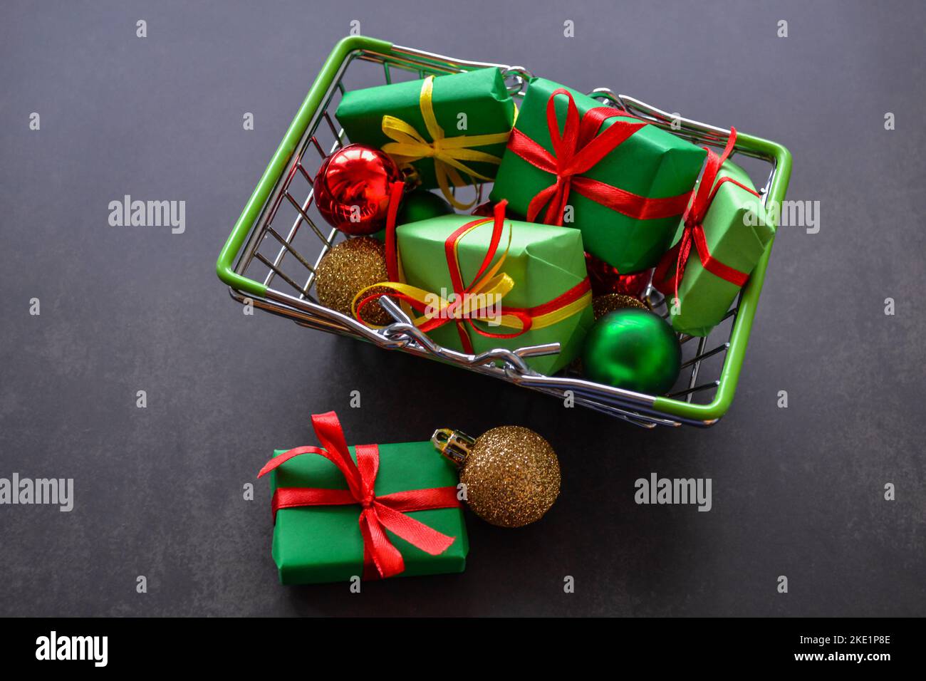 Panier avec cadeaux en papier vert, noeuds rouges et jaunes, boules de Noël en rouge, vert, or sur fond noir. Quelques cadeaux, des boules sont près. Banque D'Images