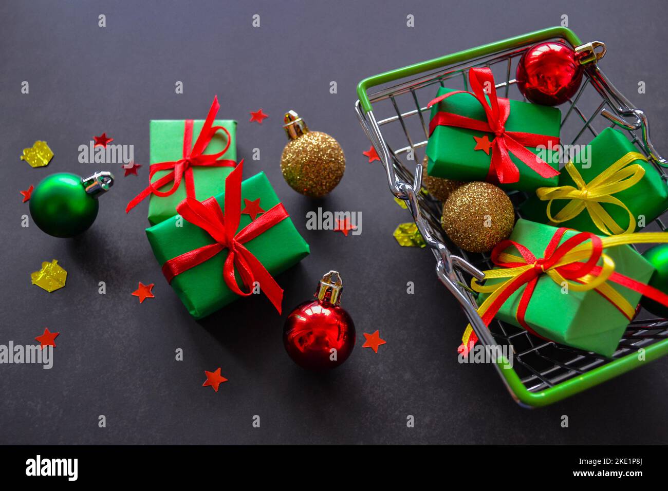 Panier avec cadeaux en papier vert, noeuds rouges et jaunes, boules de Noël en rouge, vert, or sur fond noir. Quelques cadeaux, des boules sont près. Banque D'Images