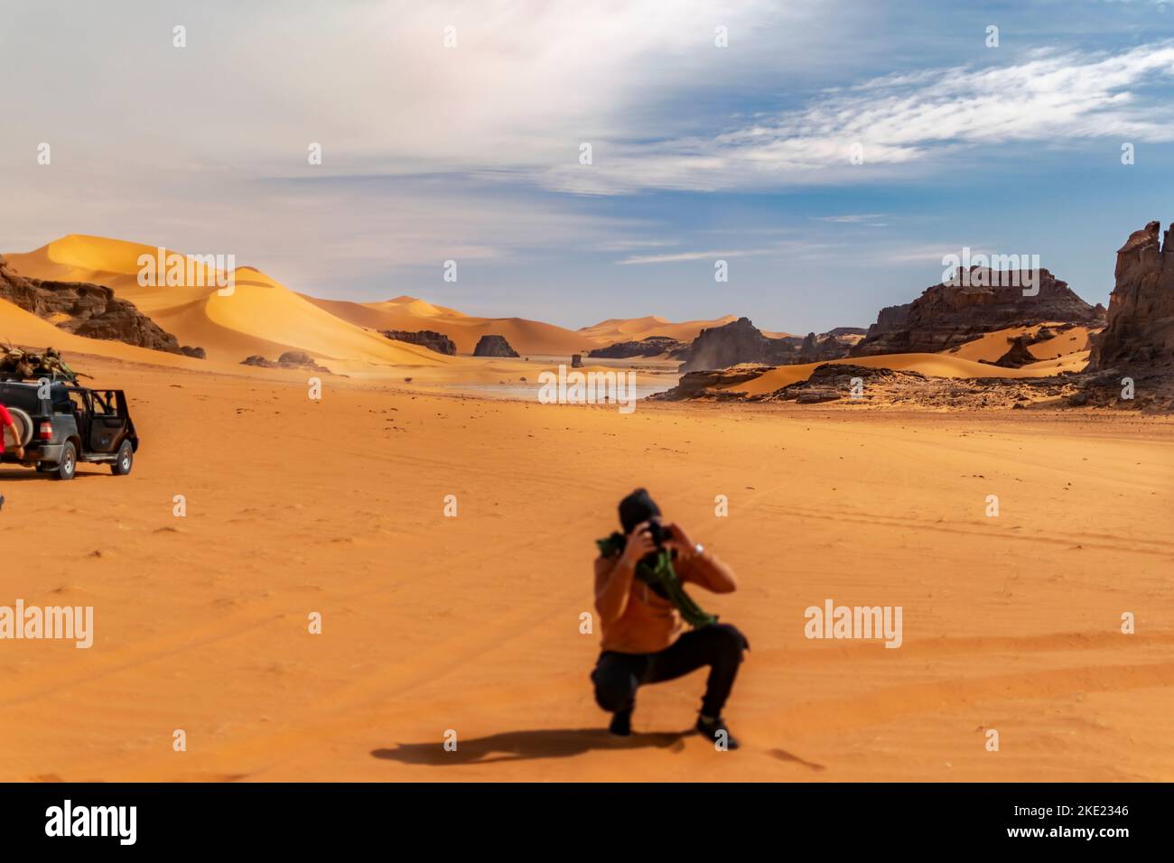 Un photographe flou se cachant et tenant un appareil photo reflex numérique. Tadrat Rouge Sahara dune de sable et montagne rocheuse. 4WD véhicule stationné sur le Banque D'Images