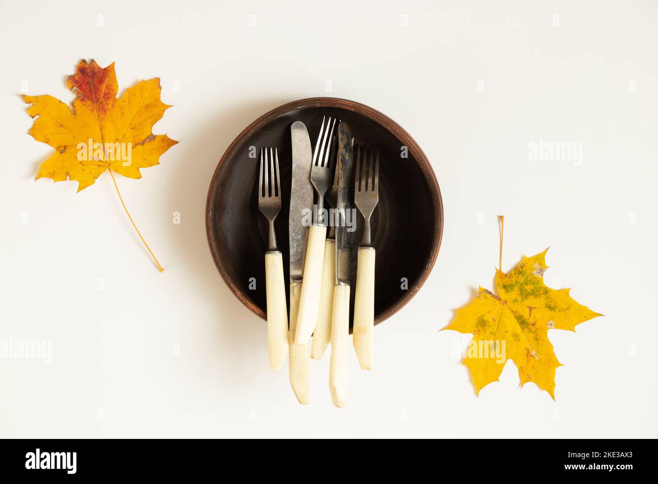 Les couteaux et les fourchettes se trouvent dans une plaque en argile profonde, qui se trouve sur un fond blanc en automne, les feuilles d'érable sèches sont en gros plan Banque D'Images
