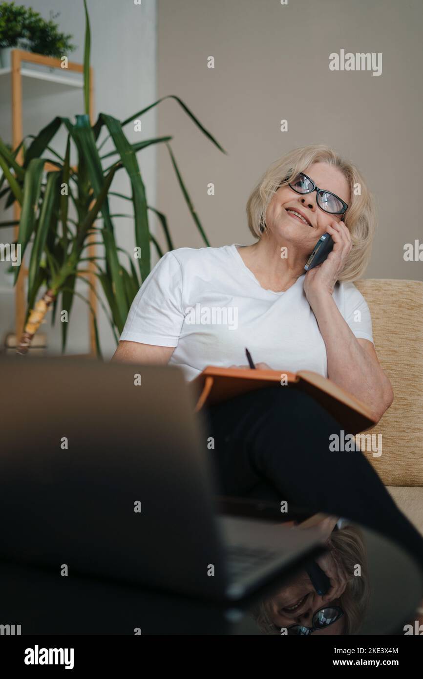 Femme âgée moderne et prospère avec lunettes et smartphone assis sur un canapé, discutant avec son ami, en faisant l'arrangement pour un dîner. Internet Banque D'Images