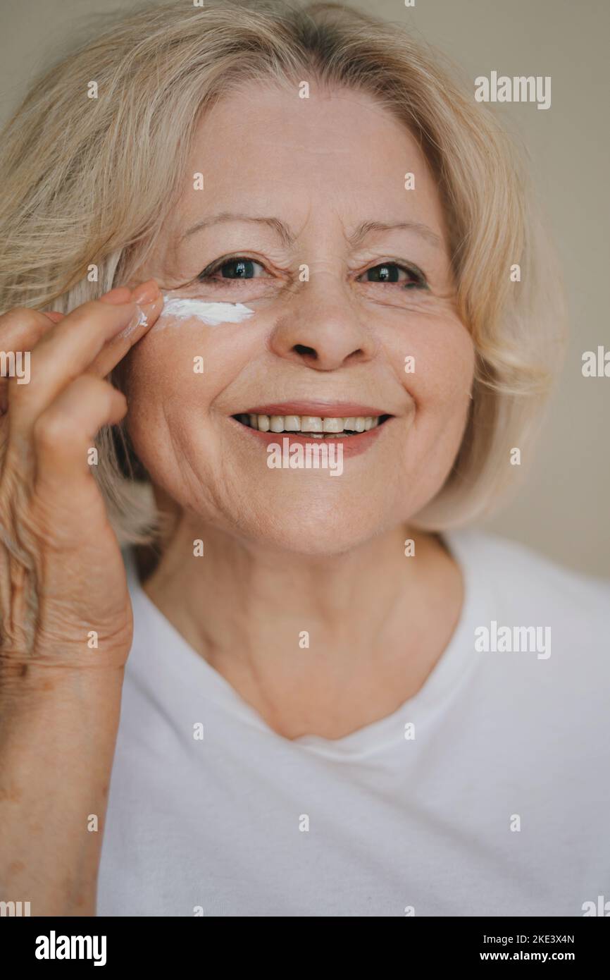 Portrait d'une femme âgée souriante appliquant une lotion anti-âge pour enlever les cercles sombres sous les yeux. Soins de la peau après 50-60 ans. Routine de beauté matinale. Banque D'Images