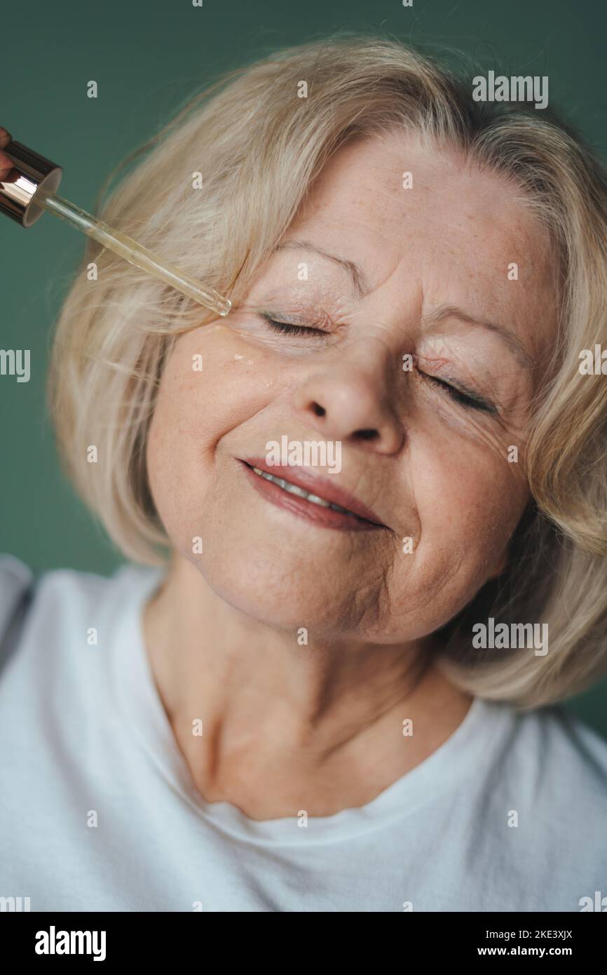 Portrait rapproché d'une femme âgée appliquant du sérum acide avec compte-gouttes sur son visage, en posant isolée sur fond vert. Femelle mature utilisant Banque D'Images