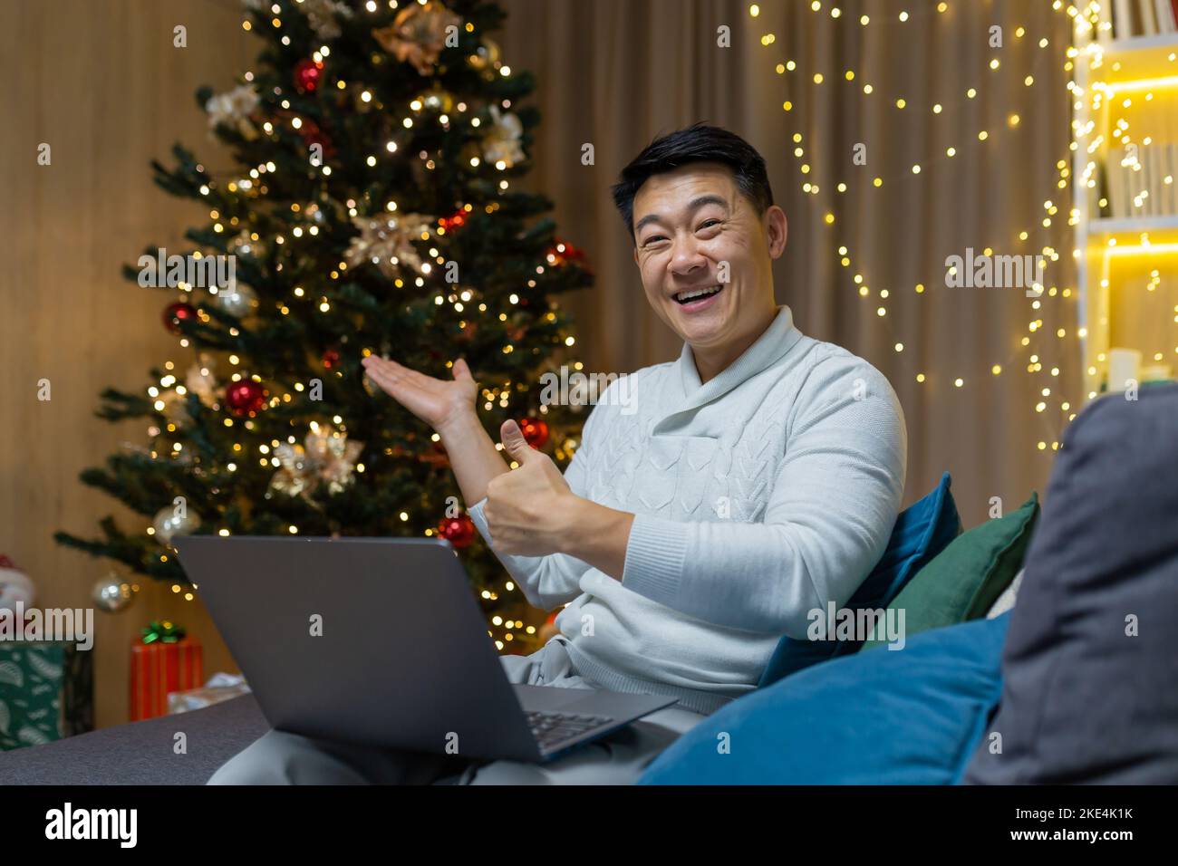 Portrait de fête d'un jeune homme asiatique beau. Assis à la maison sur le canapé près de l'arbre de Noël, tenant un ordinateur portable, regardant la caméra, souriant, pointant vers l'arbre, pointant vers le super. Banque D'Images