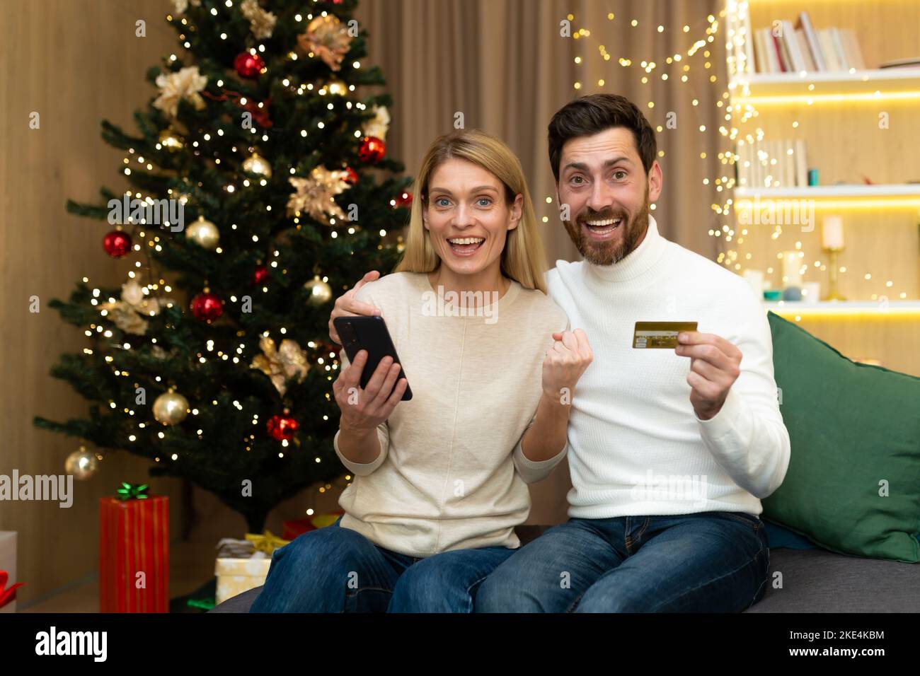 Achats en ligne. Joyeux jeune couple à la maison près de l'arbre de Noël. Un homme et une femme tiennent une carte de crédit et un téléphone dans leurs mains, acheter des cadeaux de Noël en ligne à un bon rabais. Banque D'Images