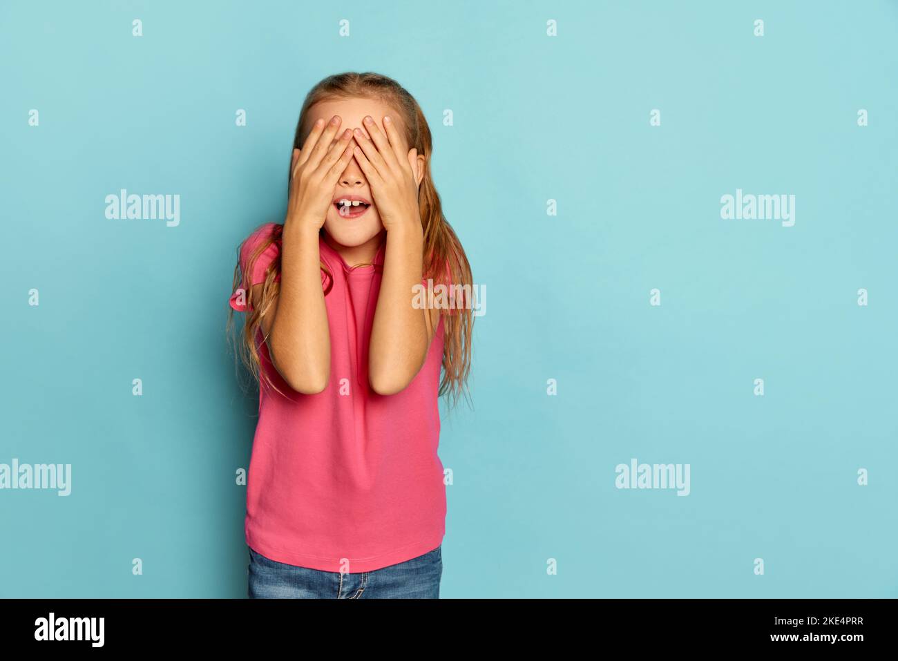 Portrait d'une petite belle fille, enfant en T-shirt rose posant avec les mains couvrant les yeux isolés sur fond bleu studio. Masquer et rechercher Banque D'Images