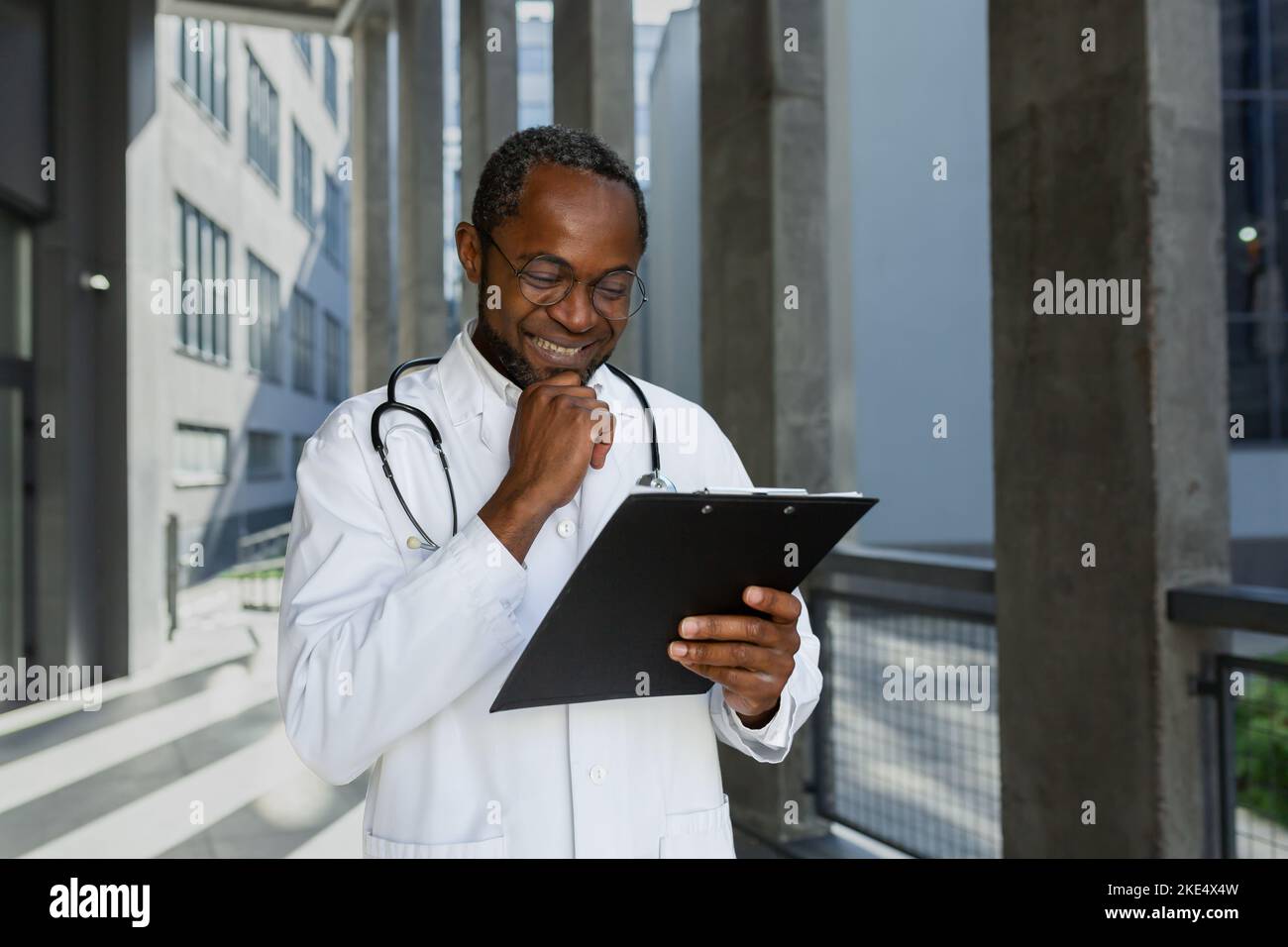 Heureux et souriant, un médecin mature lisant des documents de rapport à l'extérieur de la clinique, un homme afro-américain dans un pelage médical et un stéthoscope et des lunettes, satisfait du résultat du traitement du patient. Banque D'Images