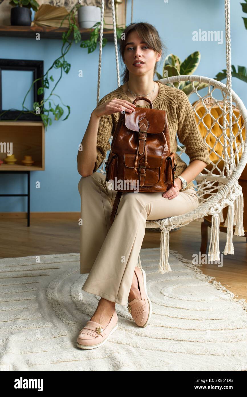 belle fille mince en pantalon beige et chandail olive posant tout en étant assis dans une chaise de balançoire avec un petit sac à dos en cuir brun brillant Banque D'Images