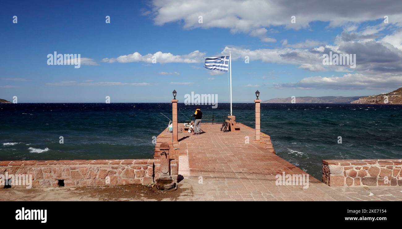 Un homme pêche avec des cannes à la jetée en pierre, le centre-ville, Petra, Lesbos. Drapeau grec soufflant dans le vent. Octobre 2022. Automne Banque D'Images