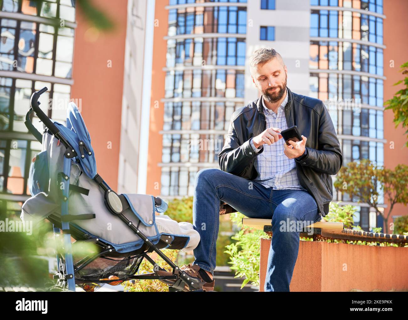Homme sérieux tenant un téléphone mobile, tout en marchant dans la rue avec un enfant en pram. Père regardant le téléphone, tout en passant du temps avec son fils dans la cour moderne des immeubles résidentiels de la ville. Banque D'Images