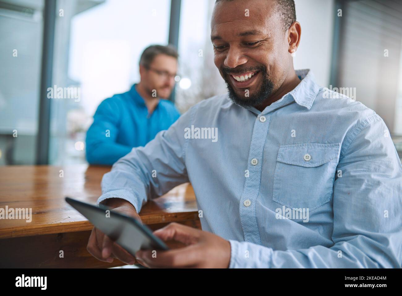 Son sourire est un signe sûr de succès. Un homme d'affaires utilisant sa tablette avec un collègue en arrière-plan. Banque D'Images