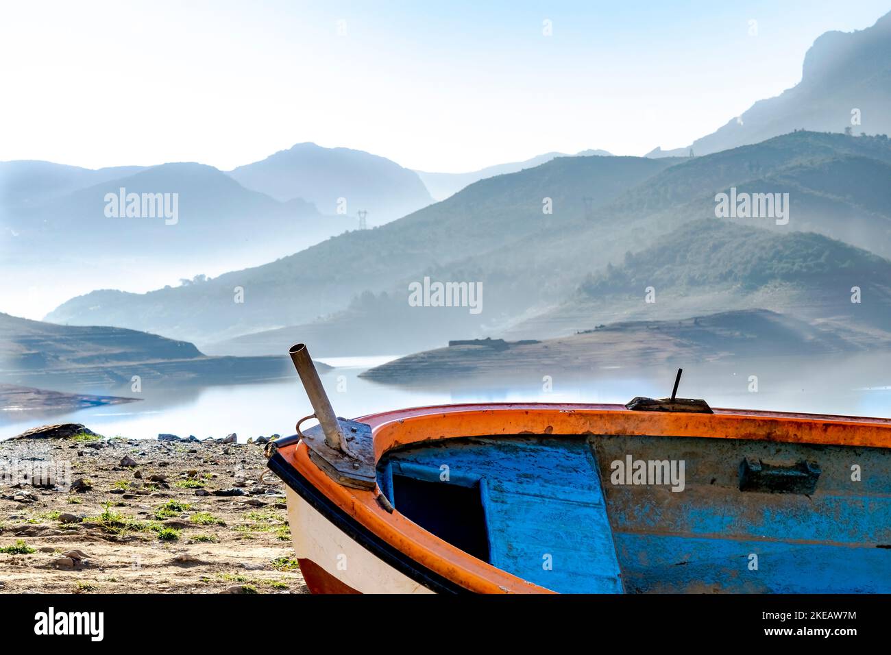 Vieux canot en bois, bateau bleu, orange et blanc coincé sur le rivage de terre sèche dans le barrage du lac de Bouzegza Keddara avec des montagnes brumeuses reflétées. Banque D'Images