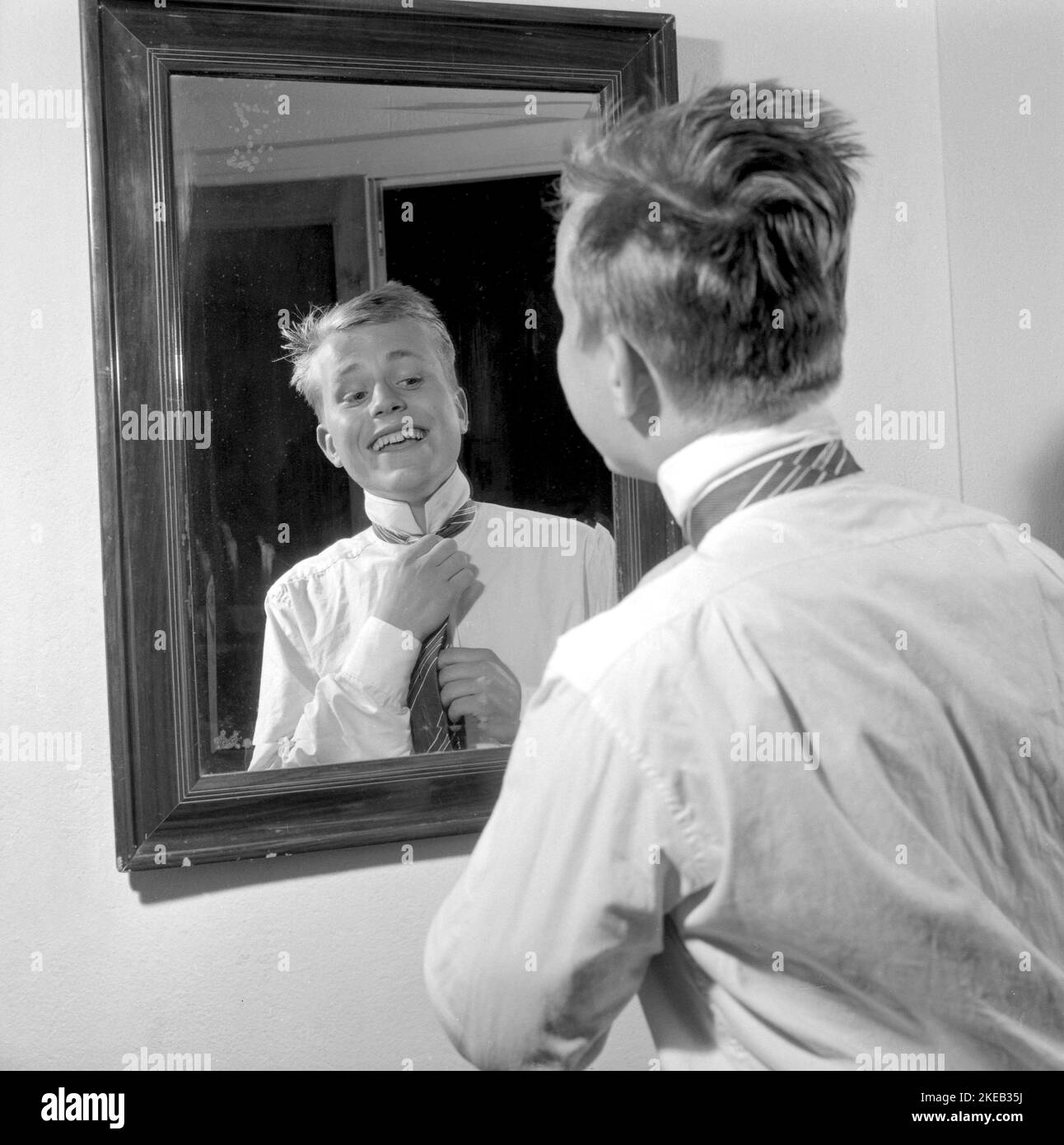 S'habiller dans le 1950s. Un jeune homme devant le miroir noue sa cravate. Il existe des variantes de nouage de la cravate, y compris l'Ascot, l'arc, le bolo, la fermeture à glissière, le cravat et la maille. Au moment où la photo a été prise, la mode et le look des cravates étaient plus minces et plus minces que les décennies précédentes. Quatre nœuds principaux sont utilisés pour nouer des cravates; un nœud à quatre mains, un nœud Pratt, un nœud à moitié Windsor et un nœud Windsor. Suède 1959. Conard réf. 4019 Banque D'Images
