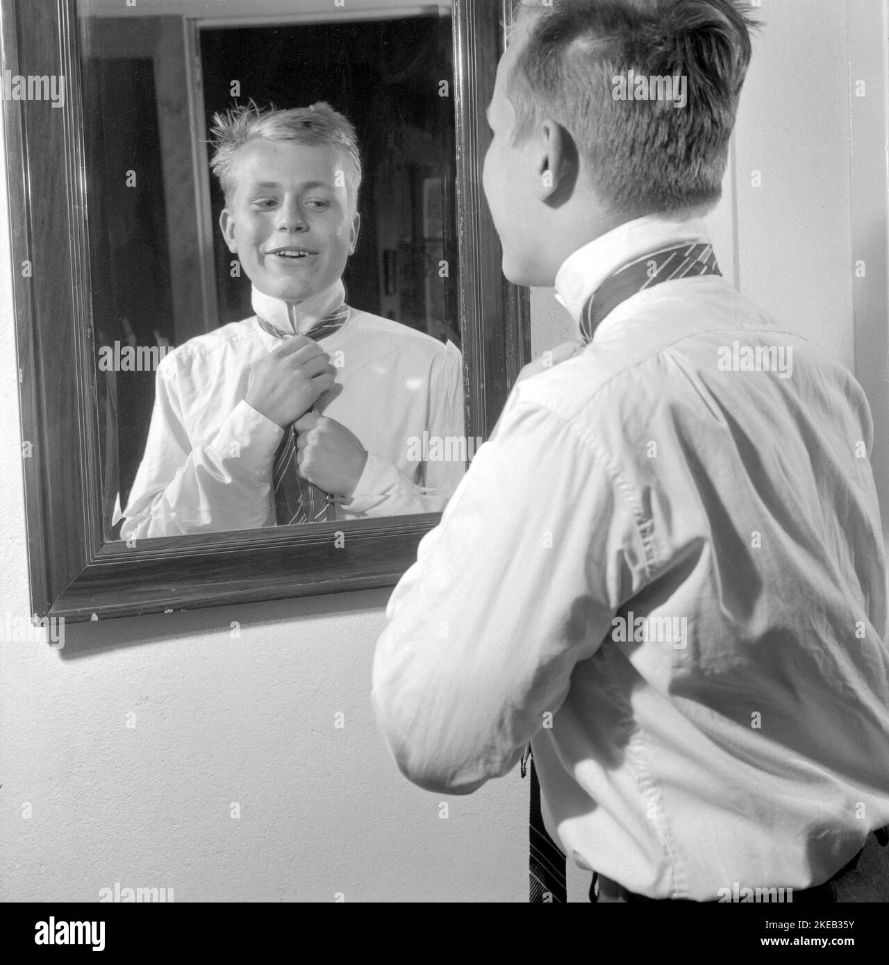 S'habiller dans le 1950s. Un jeune homme devant le miroir noue sa cravate. Il existe des variantes de nouage de la cravate, y compris l'Ascot, l'arc, le bolo, la fermeture à glissière, le cravat et la maille. Au moment où la photo a été prise, la mode et le look des cravates étaient plus minces et plus minces que les décennies précédentes. Quatre nœuds principaux sont utilisés pour nouer des cravates; un nœud à quatre mains, un nœud Pratt, un nœud à moitié Windsor et un nœud Windsor. Suède 1959. Conard réf. 4019 Banque D'Images