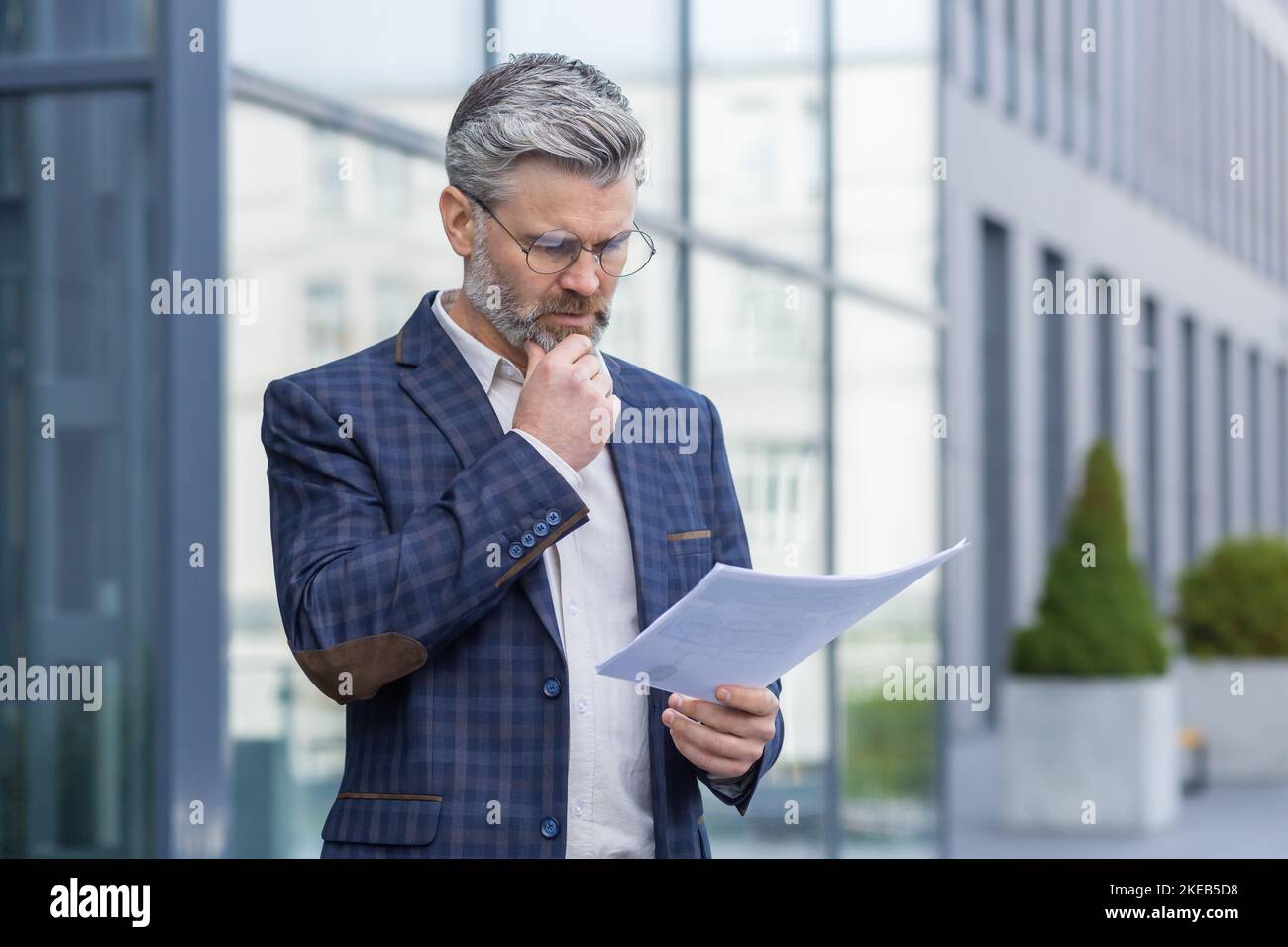 Homme d'affaires mûr à cheveux gris pensant et lisant le rapport de document financier, homme d'affaires senior en costume à l'extérieur du bâtiment de bureau avec des factures. Banque D'Images