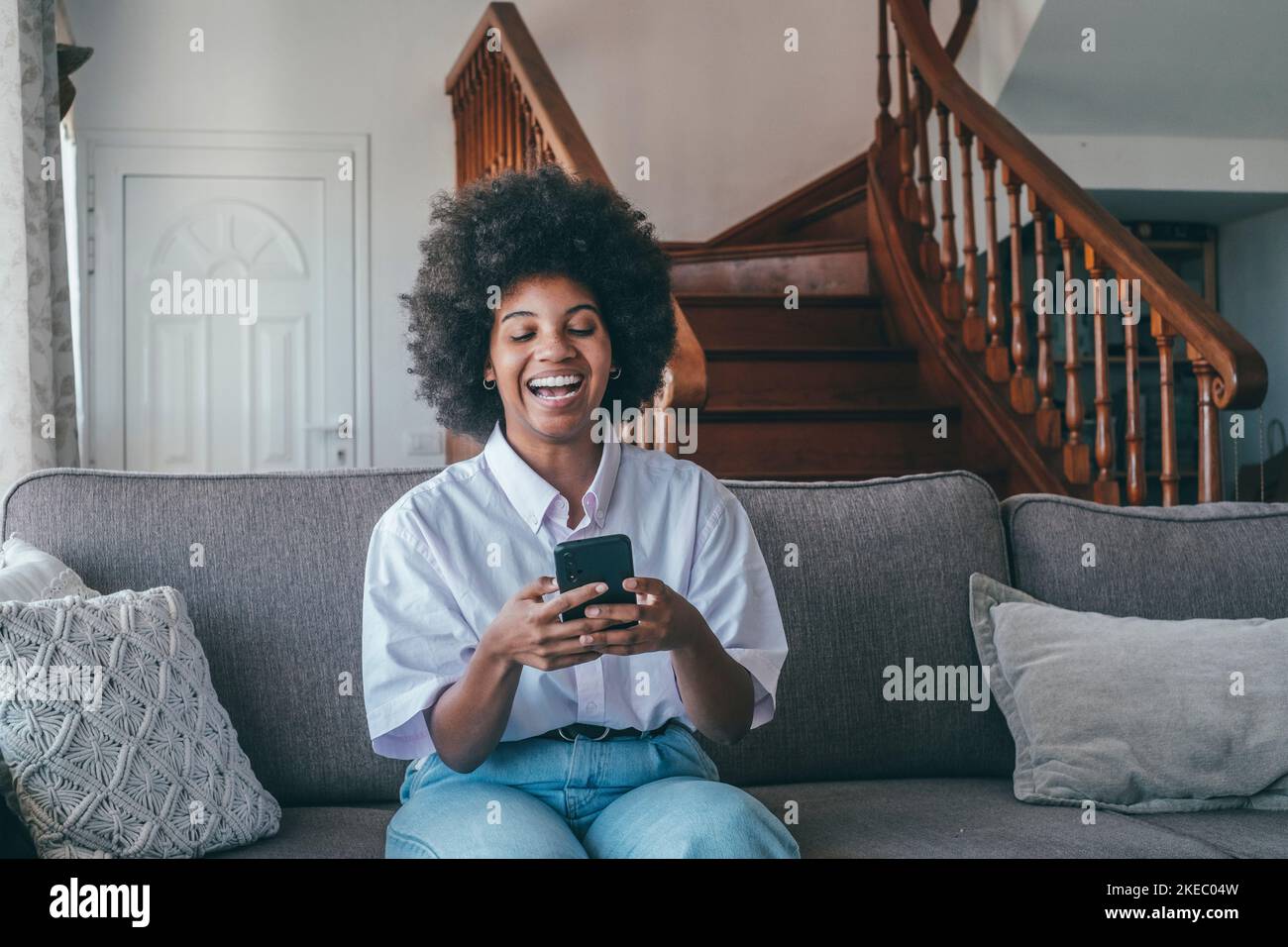 Femme afro-américaine du millénaire utilisant un téléphone portable assis sur un canapé confortable à la maison. Bonne dame noire riant à voix haute tout en lisant un SMS amusant en ligne sur smartphone Banque D'Images